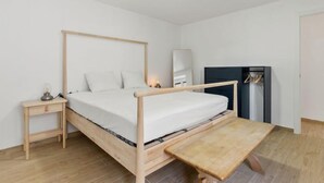 Ein schlichter Schlafraum mit einem hölzernen Bett, Nachttisch und einer Bank.