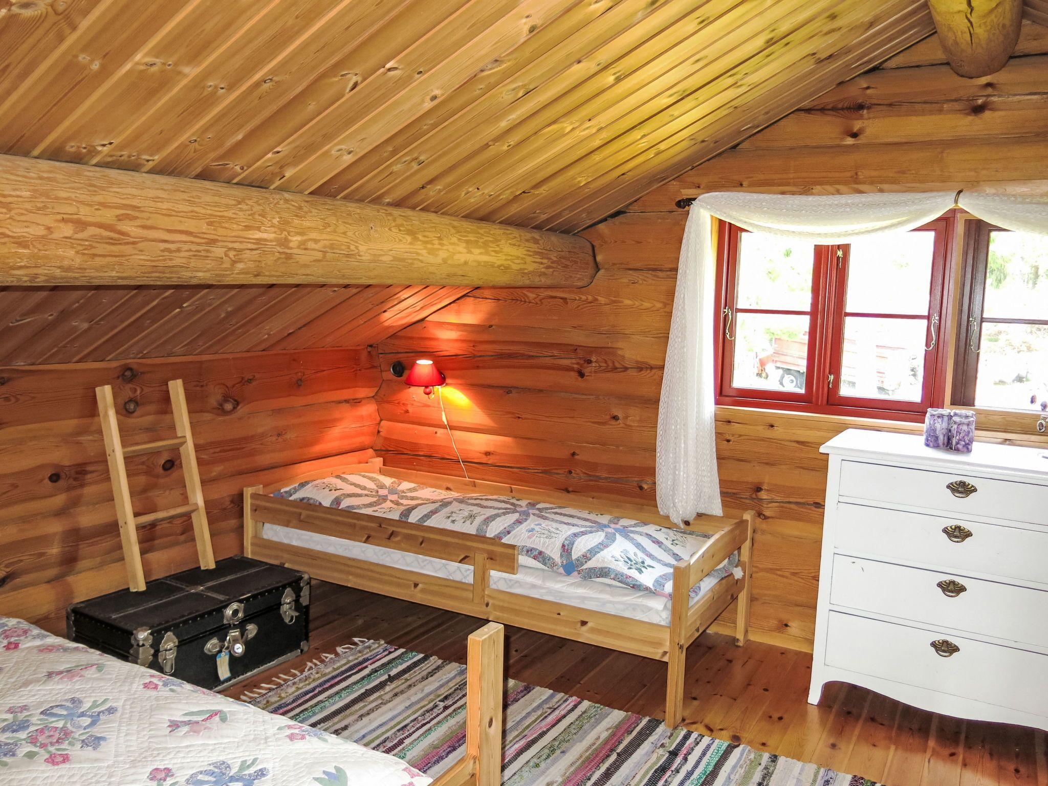 A cozy wooden cabin room with a bunk bed, a dresser, a suitcase, and a window with curtains.