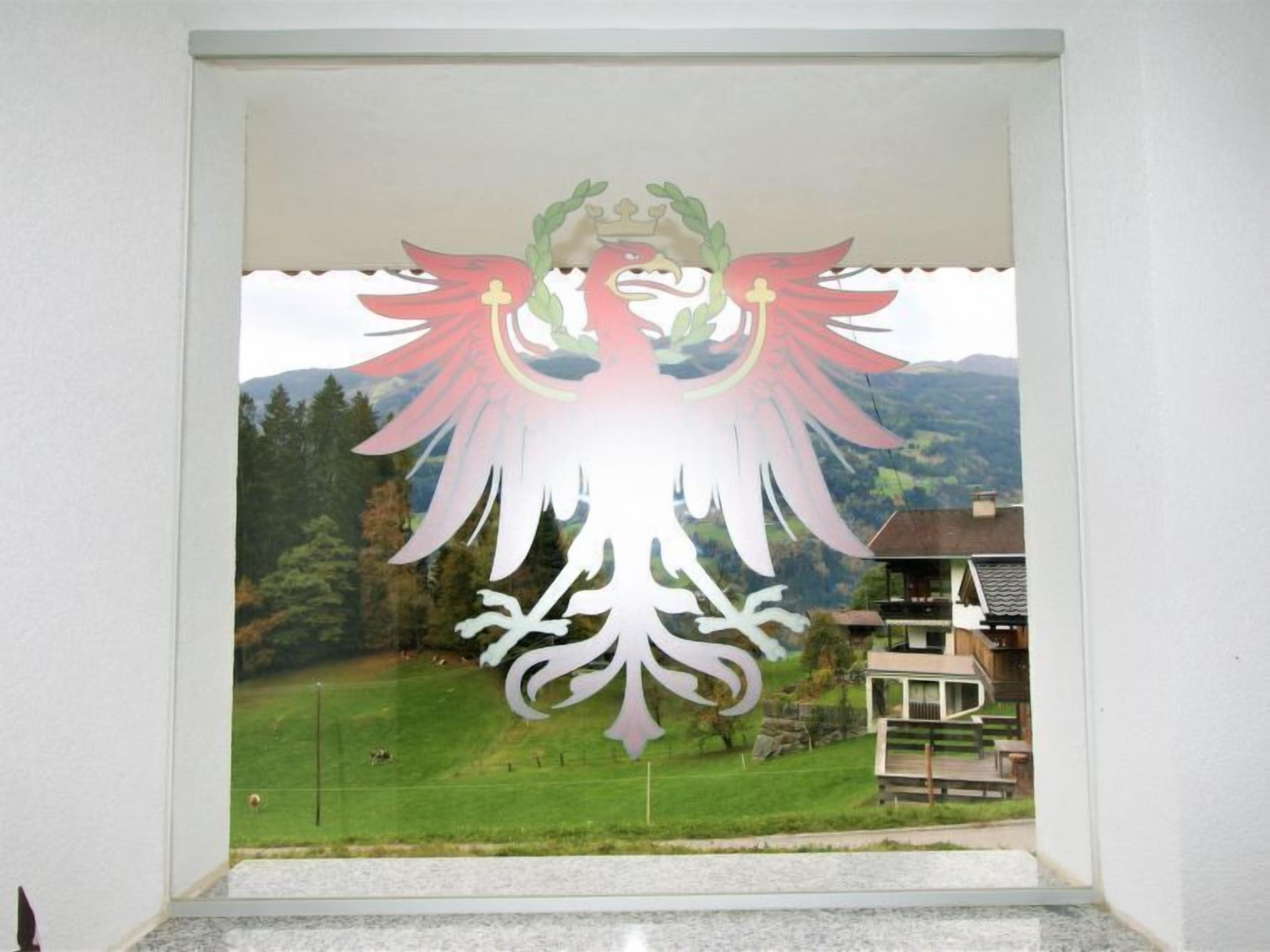 Blick durch ein Fenster mit einer verzierten Vogelornamentik, vermutlich ein Pfau, auf eine Landschaft aus grünen Feldern und Häusern im Hintergrund.