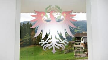 Blick durch ein Fenster mit einer verzierten Vogelornamentik, vermutlich ein Pfau, auf eine Landschaft aus grünen Feldern und Häusern im Hintergrund.