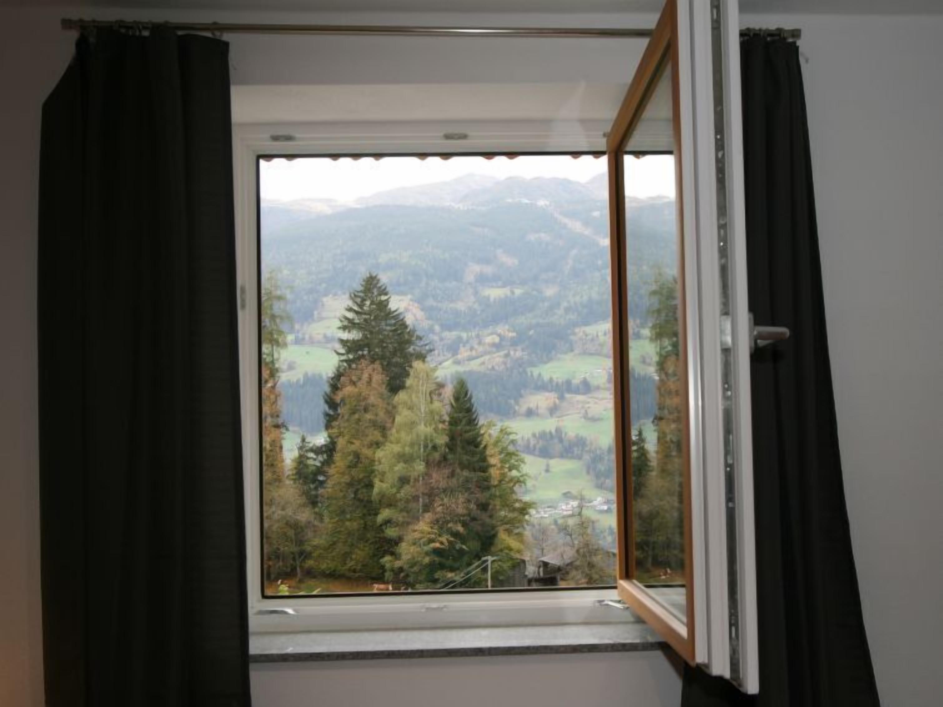 Una ventana con vistas a un paisaje montañoso, cortinas entreabiertas y una barandilla de metal.