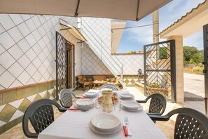 Un espace extérieur aménagé pour les repas, avec une table dressée pour quatre personnes, entourée de chaises, et une vue sur un bâtiment à la façade recouverte de carreaux.