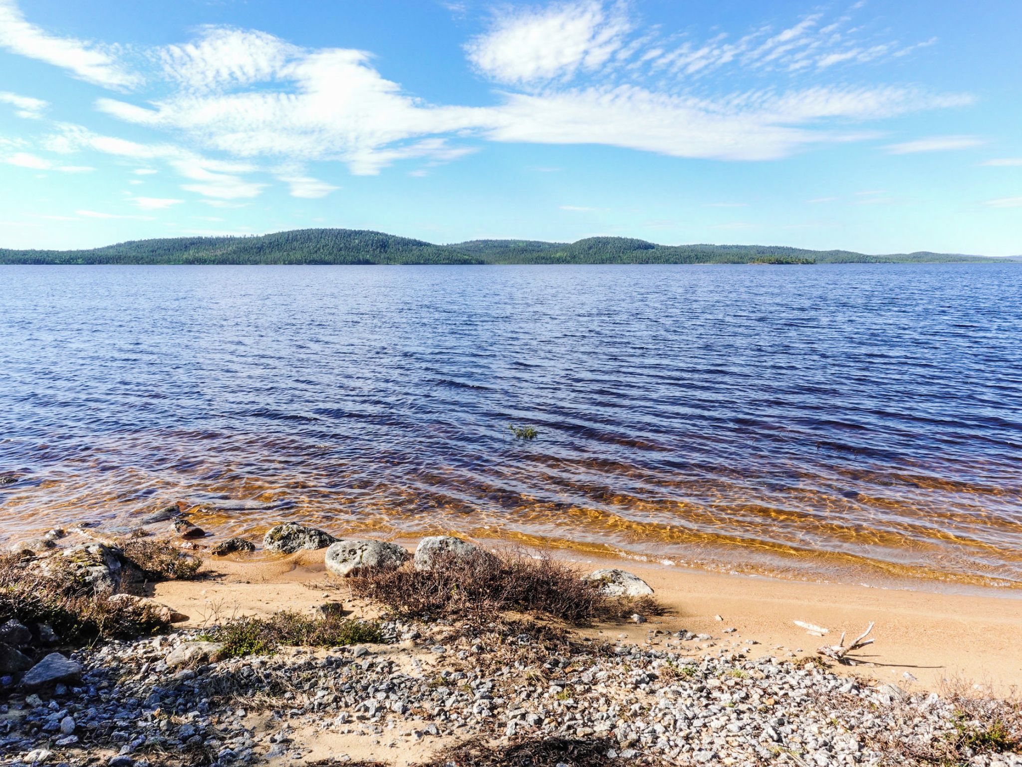 Une plage à l’eau claire, bordée de galets, avec des collines qui se dessinent à l’horizon.