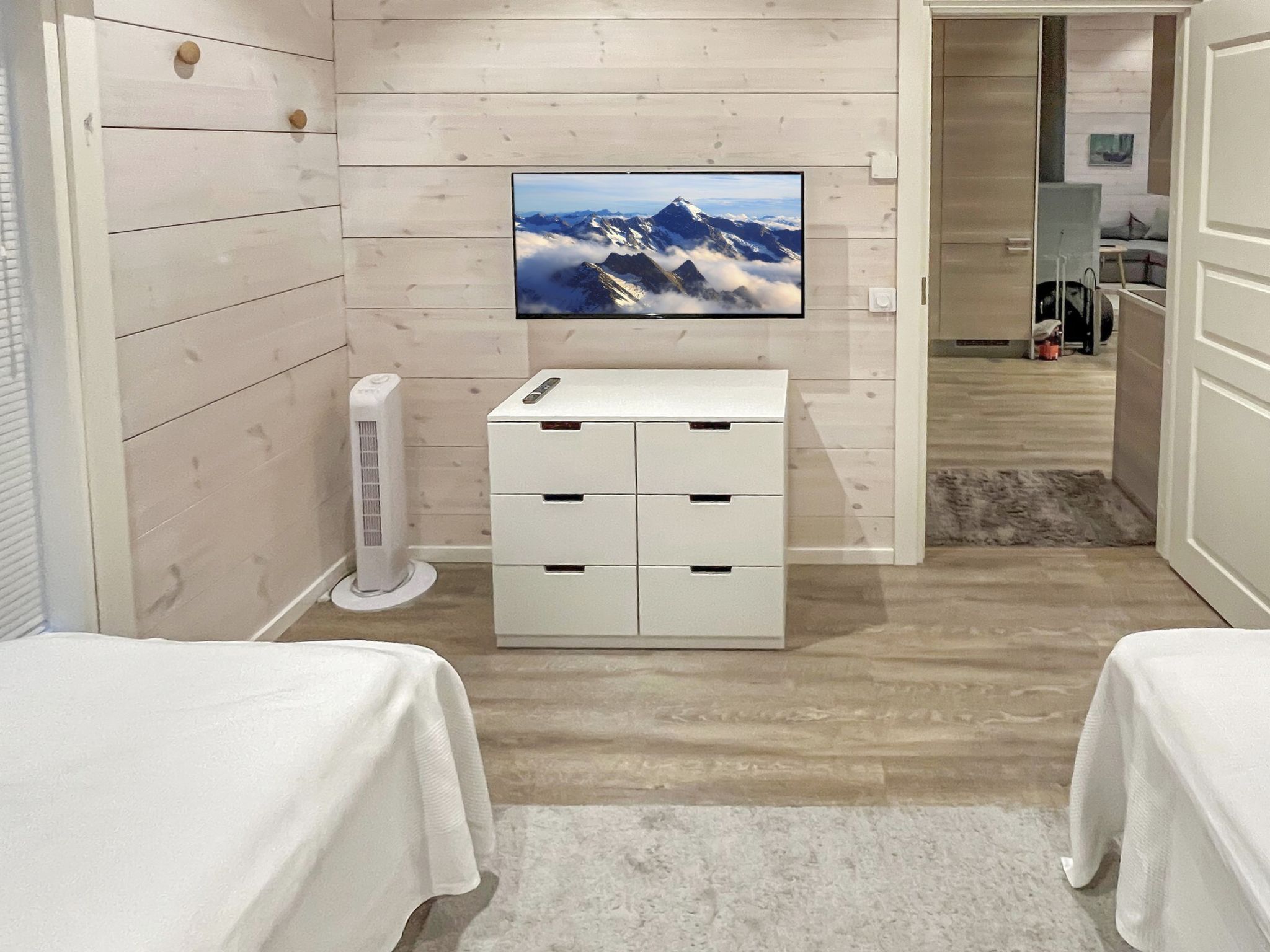 Habitación de hotel con una televisión instalada en la pared, un equipo de aire acondicionado blanco, un mueble blanco para guardar objetos y dos camas con ropa blanca.
