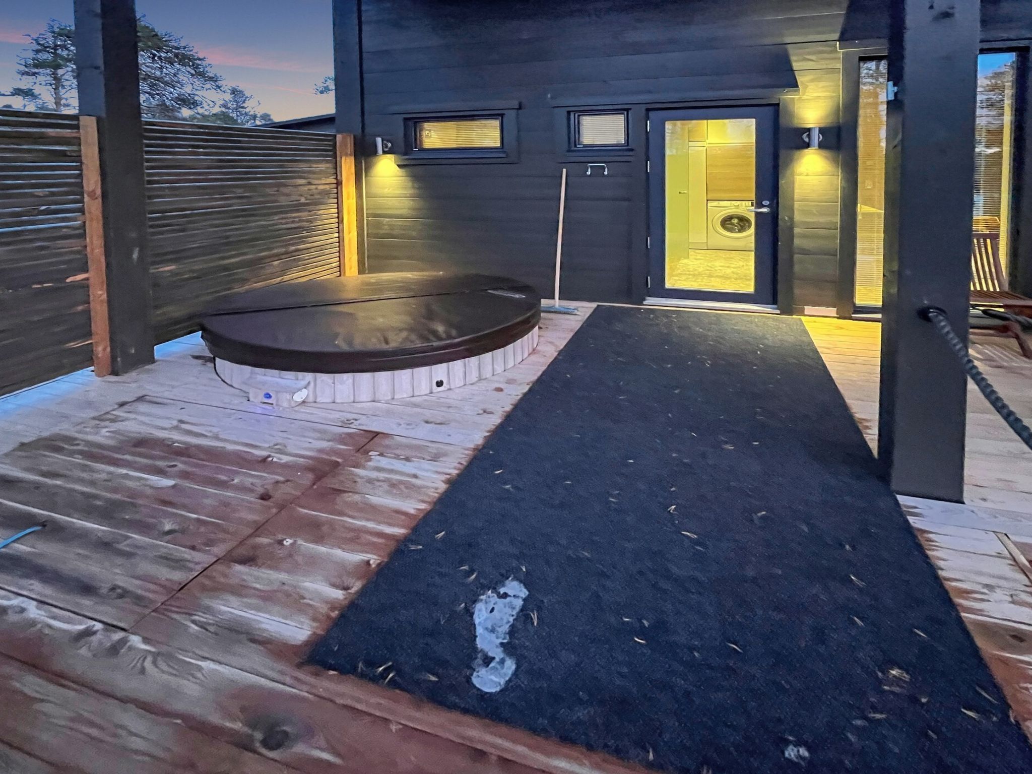 Area esterna coperta con un pavimento in legno, una vasca idromassaggio circolare e un tappetino in gomma nera che conduce a una porta.