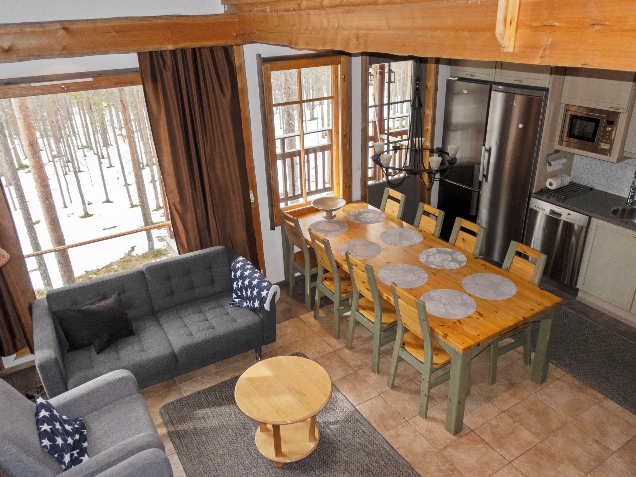 Un intérieur chaleureux avec une table à manger dressée pour six personnes, un canapé, une petite table ronde et un coin cuisine équipé d’appareils électroménagers modernes.
