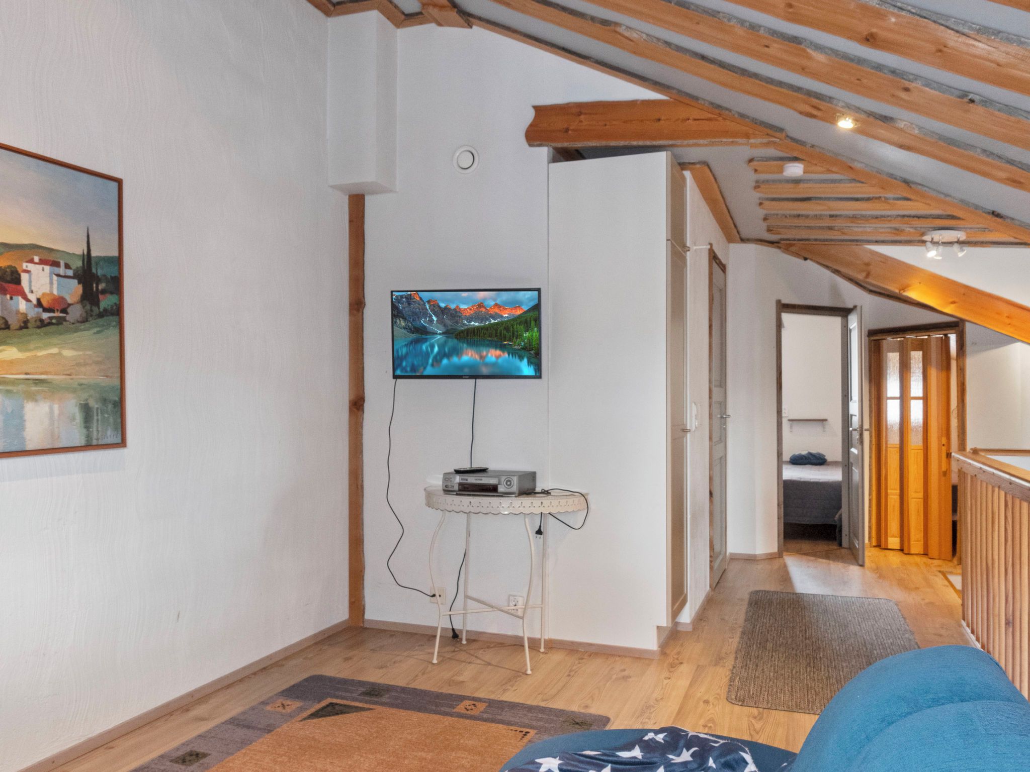Una habitación con una televisión montada en la pared, una mesita con un dispositivo encima, suelo de madera y una puerta que da a otra habitación.