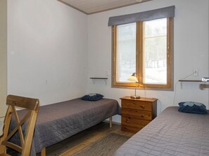 A small, simple room with two single beds, a wooden chair, a dresser, and a window with blinds.