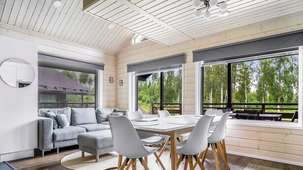 Un interior moderno con techo de madera, una mesa redonda de comedor con sillas, un sofá gris y ventanas amplias con vistas a árboles.