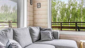 A grey sectional sofa with cushions, a wooden deck, and a view of trees through a window.