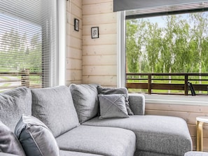 Un sofá seccional gris con cojines, una terraza de madera y vista a árboles a través de una ventana.