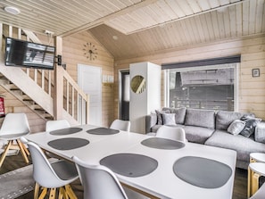 Un interior moderno con una mesa y sillas blancas, un sofá gris, un televisor montado en la pared y una escalera.
