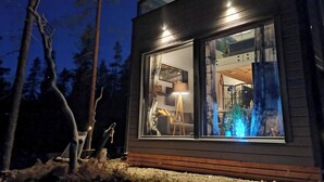 A modern house with a large glass window, wooden exterior, and a visible interior with a lamp and plants. - Levi secret igloo revontuli 10d by Interhome (Kittilä)