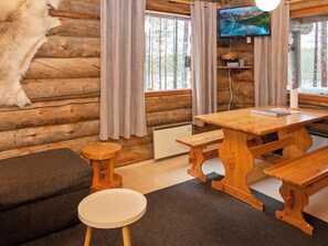 Interior rústico de una cabaña con una mesa de madera, bancos, un sofá y un televisor montado en la pared.