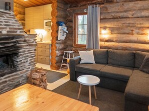 Interior acogedor de una cabaña de troncos con chimenea de piedra, un sofá, una pequeña mesa redonda y una zona de cocina con microondas.