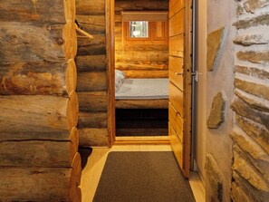 Interior de una cabaña de troncos con una cama, paredes de madera y un ventanal pequeño.