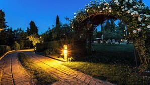 Ein gepflasterter Weg führt zu einem gut beleuchteten Torbogen, im Hintergrund befinden sich ein Garten und Bäume.