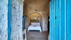 Un étroit couloir en pierre mène à une pièce où se trouvent un lit, un téléviseur et une porte bleue.