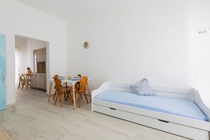 Ein Zimmer mit einem weißen Bett, einem Esstisch mit Stühlen und einer Küchenzeile im Hintergrund.