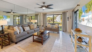Ein geräumiges Wohnzimmer mit grauem Sofa, einem Couchtisch und einem Fernseher. Es gibt Deckenventilatoren, ein großes Fenster mit Blick auf Palmen und einen Balkon mit Geländer.