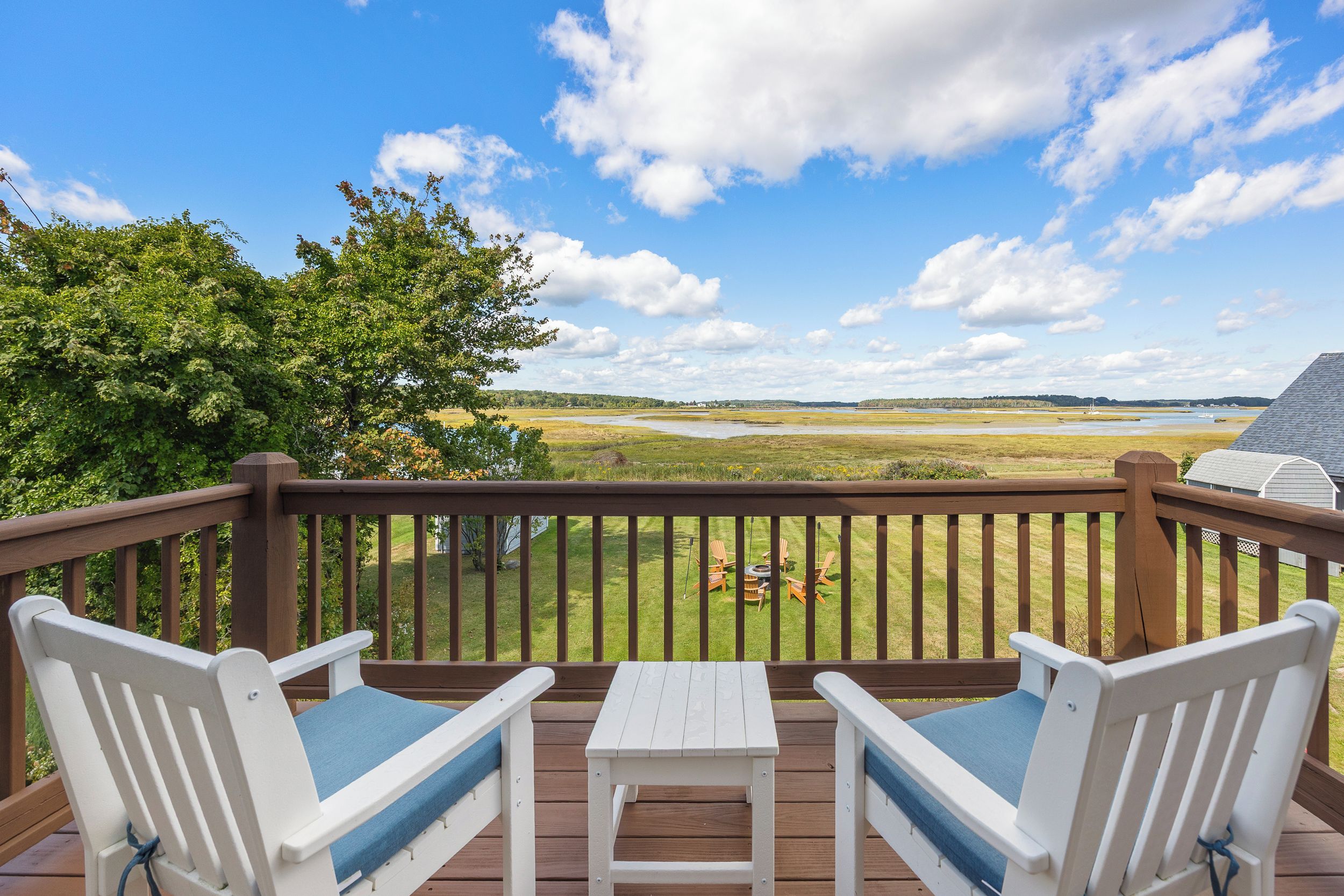 A deck with two white chairs and a table, overlooking a grassy landscape with a body of water and trees.