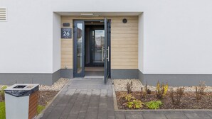A modern building entrance with a glass door, a number sign '26', and a small garden area with plants.