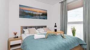 A bedroom with a bed, a bedside table with a lamp, a window with curtains, and a painting on the wall.