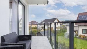 A balcony with a wicker sofa, a glass railing, and a view of neighboring houses and a garden.