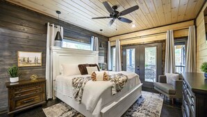 A bedroom with a wooden ceiling, a bed with white bedding, a nightstand, a chair, and a view of the outdoors. - Greyhawk Pines|Sleeps16|HotTub|Arcade|Shuffleboard (Broken Bow)