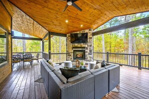 A covered outdoor seating area with a stone fireplace, wooden deck, and a view of the surrounding forest.