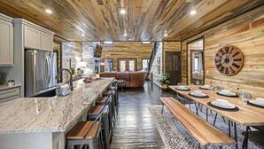 A modern kitchen with a large island, wooden ceiling, and a dining area with a wooden table and benches. - Greyhawk Pines|Sleeps16|HotTub|Arcade|Shuffleboard (Broken Bow)