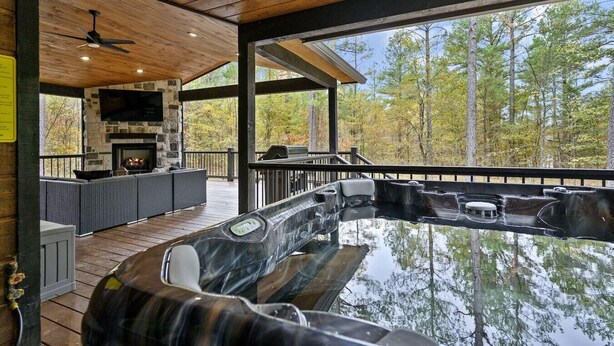 A hot tub area with a stone fireplace, outdoor seating, and a view of trees. - Greyhawk Pines|Sleeps16|HotTub|Arcade|Shuffleboard (Broken Bow)