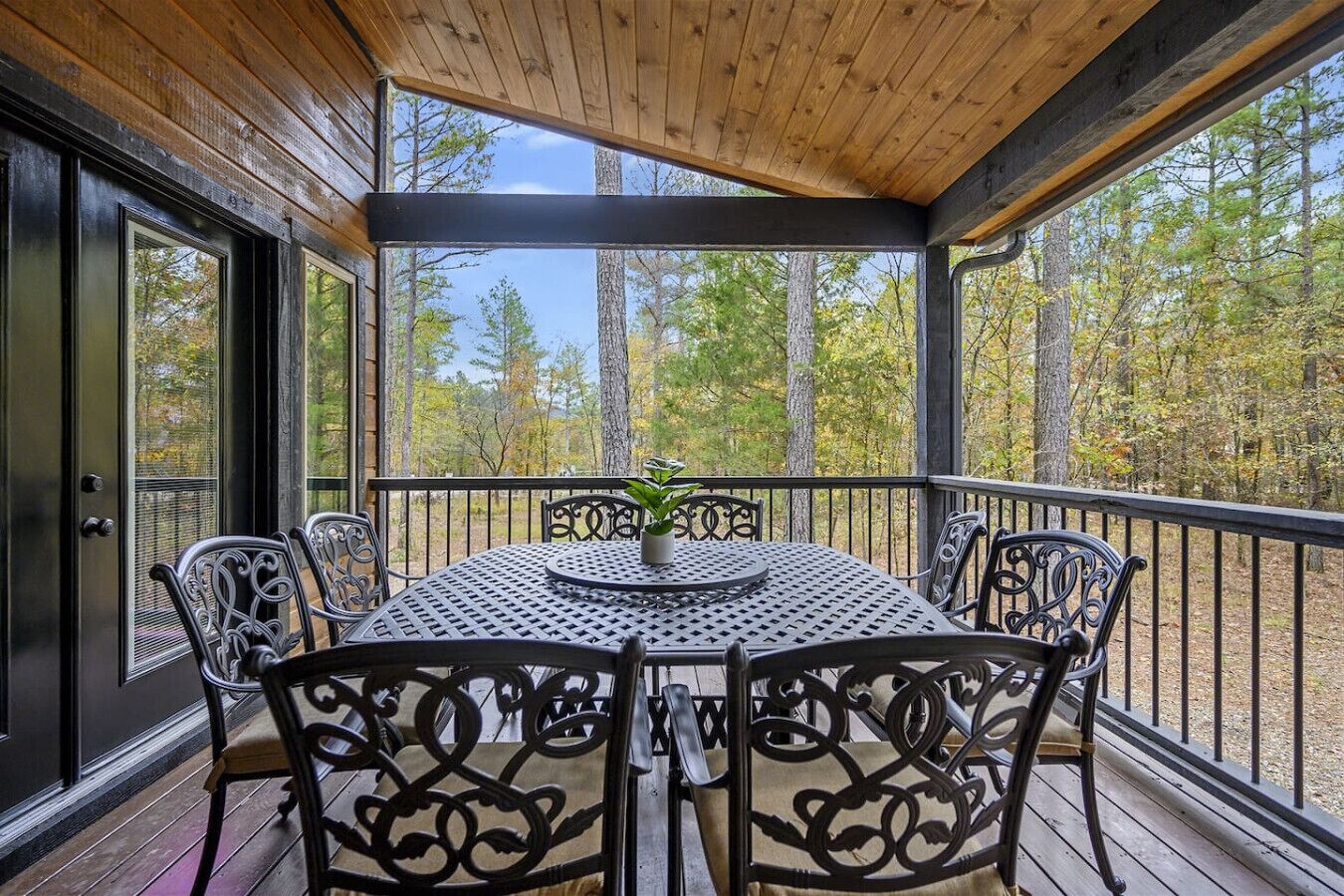 A covered outdoor seating area with a round table and chairs, surrounded by a wooden deck and metal railings, overlooking a forested area.