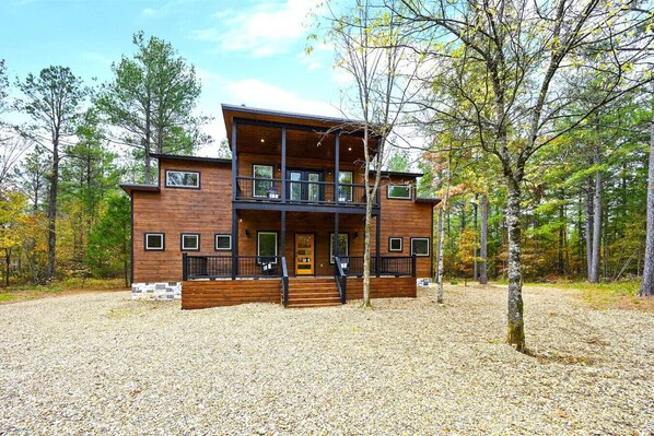 A two-story wooden cabin with a balcony, surrounded by trees and gravel.