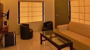 A modern living room with a sofa, a glass coffee table, and two black ottomans. There are two windows with blinds, a flat-screen TV mounted on the wall, and a tiled floor. - The Windy Mist Resort Munnar (Udumbanchola)