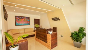 A reception area with a wooden reception desk, a sofa, a painting, and potted plants. - The Windy Mist Resort Munnar (Udumbanchola)