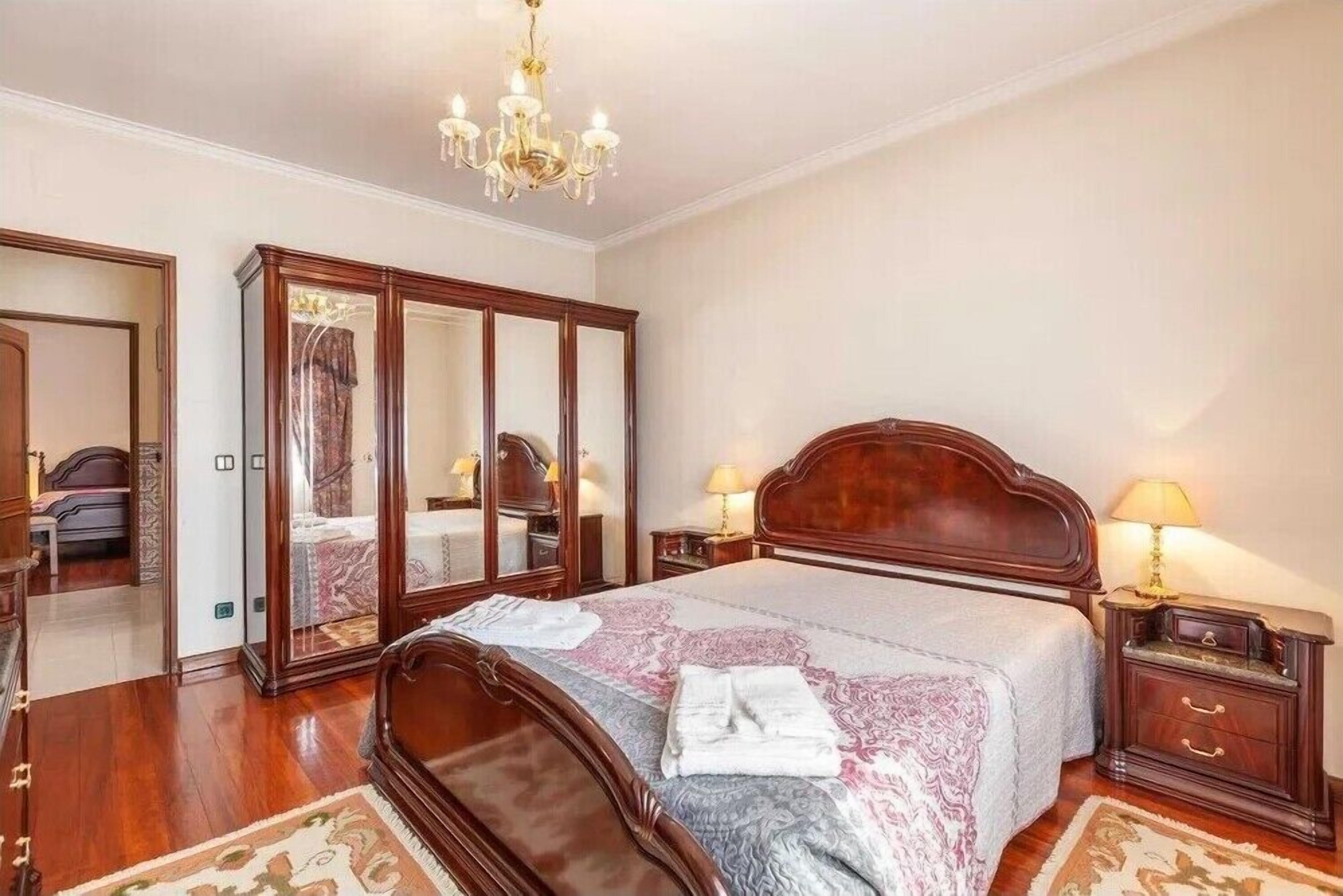 A bedroom with a large bed, wooden headboard, two bedside tables with lamps, a chandelier, and a large mirror.