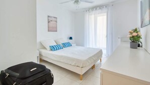 Une chambre à coucher comprenant un lit, un ventilateur de plafond, une table de chevet, une lampe, une fenêtre avec des rideaux et une plante en pot.