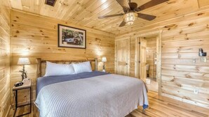 Un dormitorio de cabaña de madera con cama, dos lámparas de noche, ventilador de techo y un cuadro enmarcado en la pared.