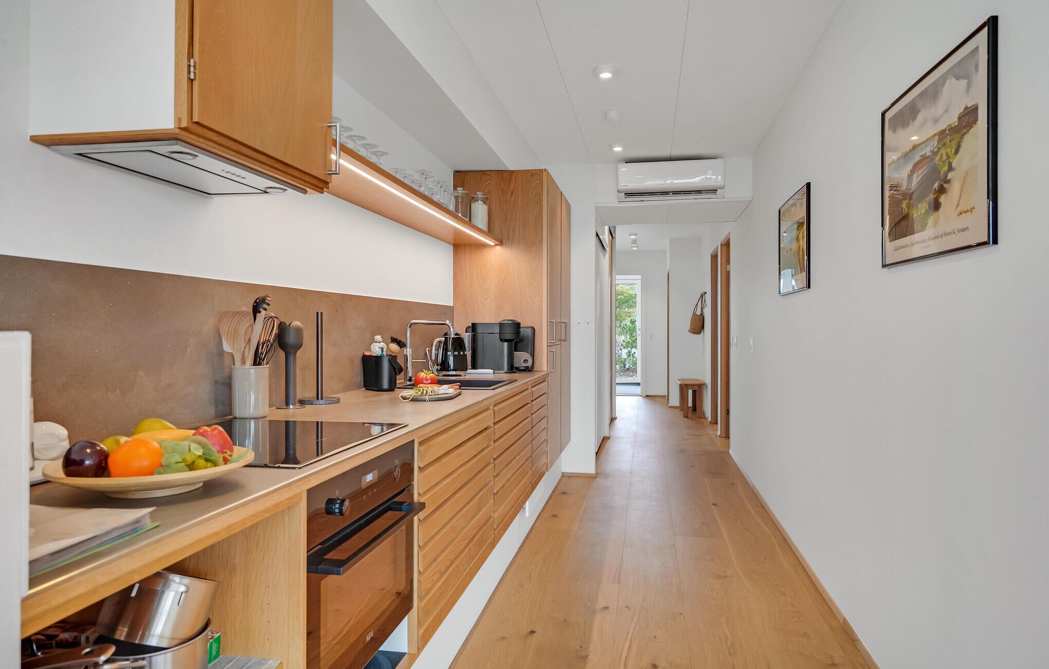 A modern kitchen with wooden cabinets, a stainless steel range hood, and a built-in oven. There is a countertop with a coffee maker and a bowl of fruit. The kitchen leads to a hallway with a view of the outdoors.