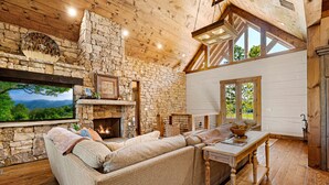 A cozy living room with a stone fireplace, wooden beams, and a large window overlooking a scenic landscape. - Copperline Lodge - located in Blue Ridge, GA (Epworth)