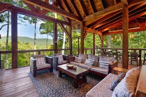Una terraza de madera al aire libre con un sofá, sillas y una mesa, con vistas a un paisaje verde exuberante.