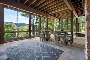 A covered outdoor area with stone flooring, wooden tables and chairs, and large windows offering a view of a forested landscape.