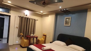 A hotel room with a bed, a desk, two chairs, a small table, a ceiling fan, and a framed picture on the wall. - Hotel Pavan Mansion (Raichur)