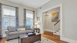 Ein Wohnzimmer mit grauem Sofa, einem dunkelholznen Couchtisch und einem gemusterten Teppich. Zwei Fenster mit Jalousien und Vorhängen, eine Lampe auf einem Beistelltisch sowie eine Treppe, die in einen anderen Raum führt.