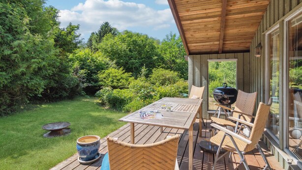 A wooden deck with a table and chairs, a grill, and a view of a lush garden.