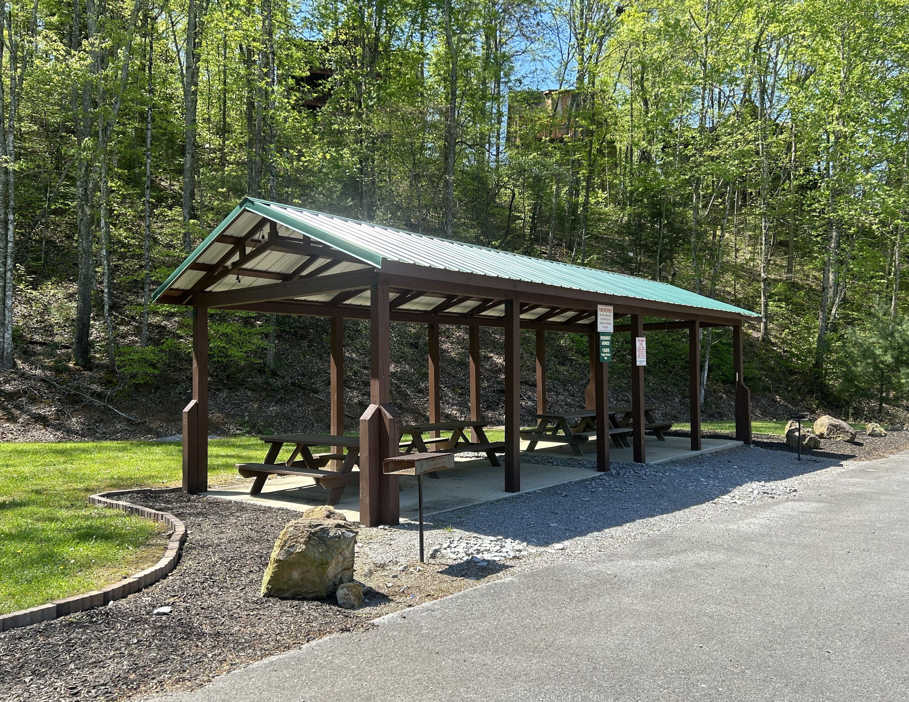 A wooden pavilion with picnic tables, surrounded by a gravel path and a grassy area.