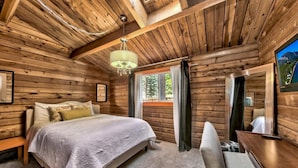 Een slaapkamer in een blokhut met een bed, een bankje, een tafel en een raam met gordijnen.