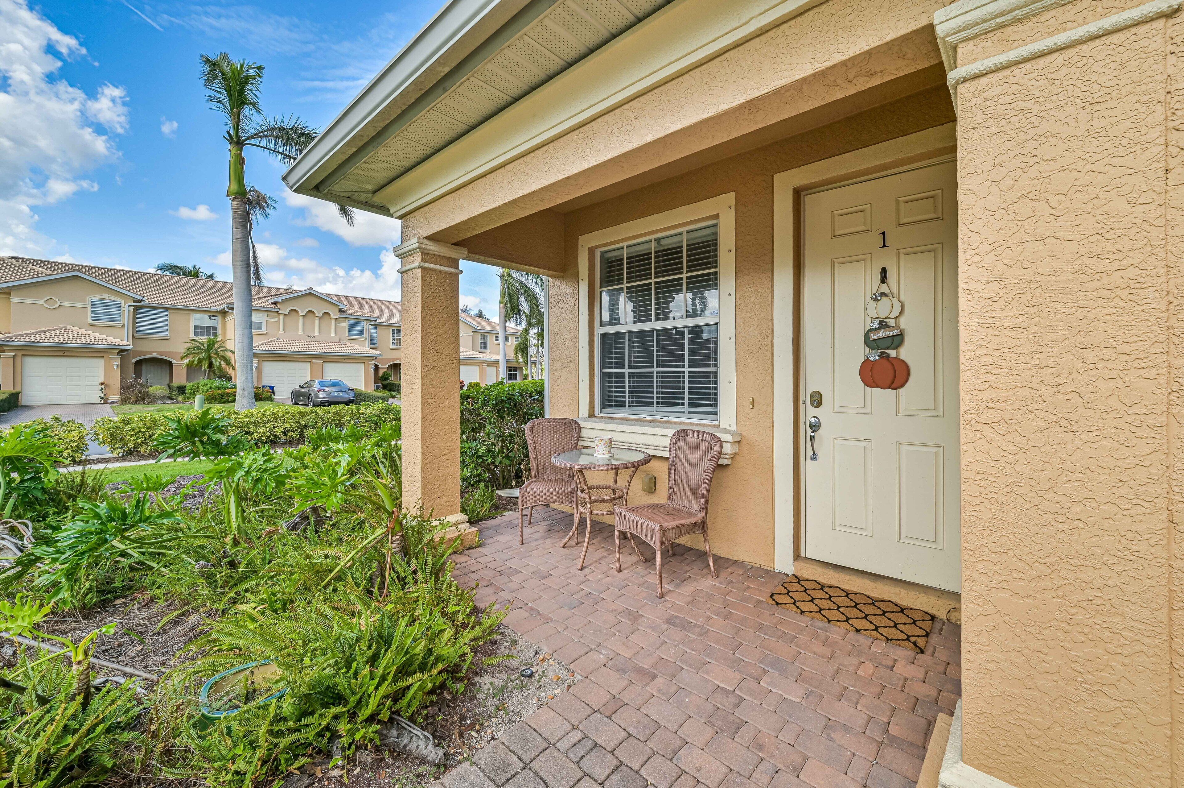 A residential property with a patio, two wicker chairs, a small table, a door with a number, and a palm tree.