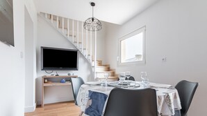 Un coin repas avec une table dressée pour quatre personnes, un téléviseur posé sur un meuble, et un escalier en arrière-plan.